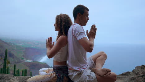 Against-the-ocean's-panoramic-view,-a-man-and-woman-sit-on-a-mountain's-rock-back-to-back,-immersing-themselves-in-meditation-and-yoga